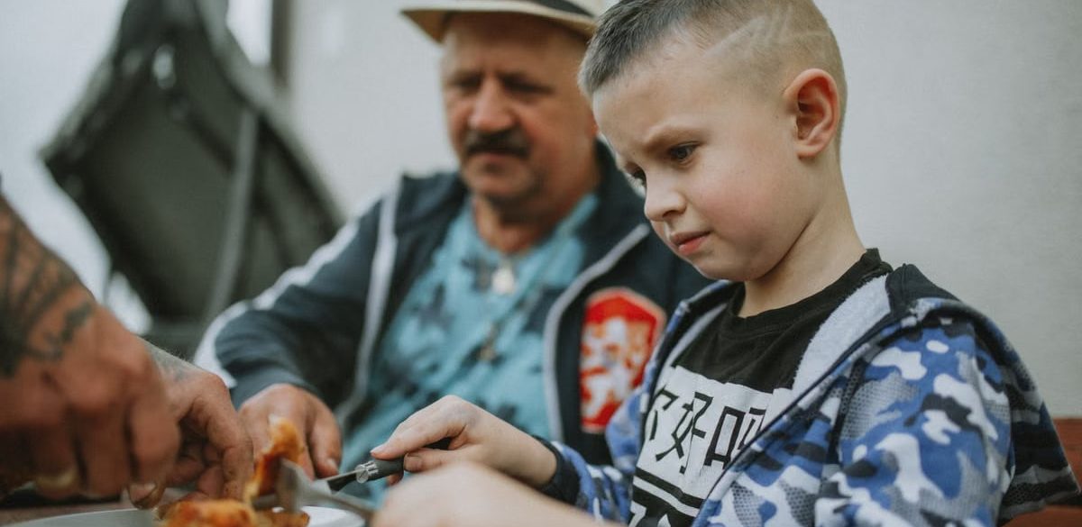 A child and his father share a meal outdoors, enjoying grilled food together.