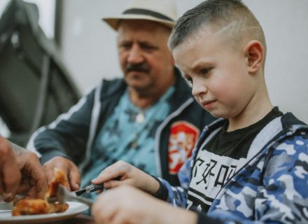 A child and his father share a meal outdoors, enjoying grilled food together.